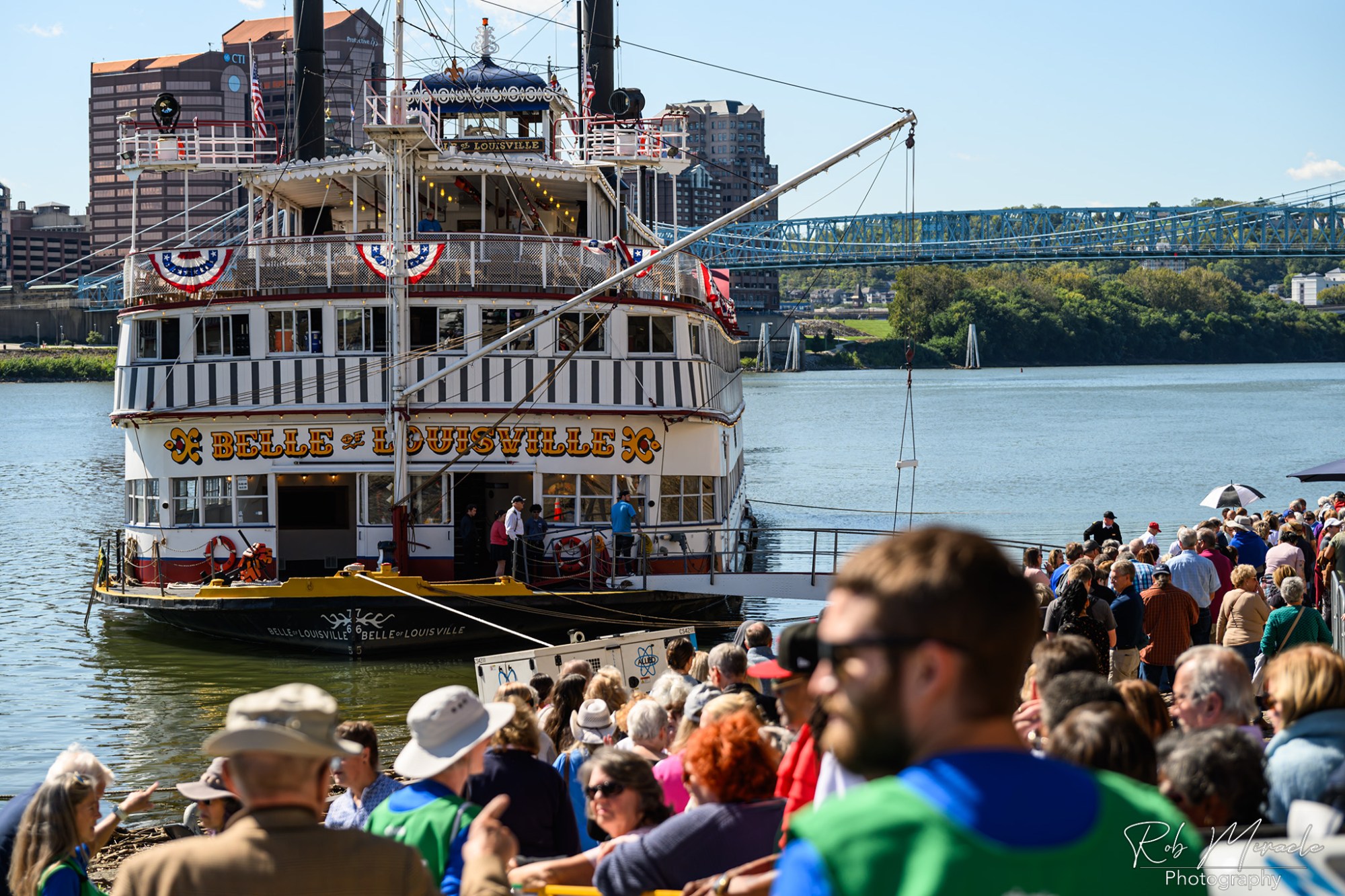 RiverRoots07_WEB Crowd boarding the 'Belle of Louisville' steamboat docked by a river on a sunny day.