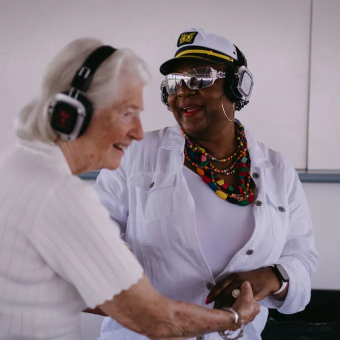 Silent Disco 1 Two women wearing headphones and smiling while holding hands in a light room.