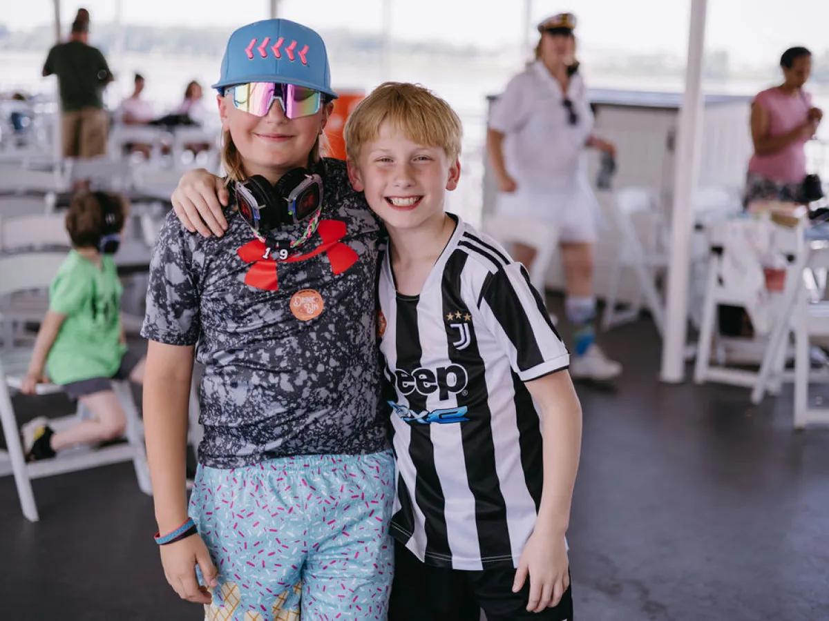 Two kids smiling, one wearing sunglasses and a cap, the other in a striped sports jersey, indoors.