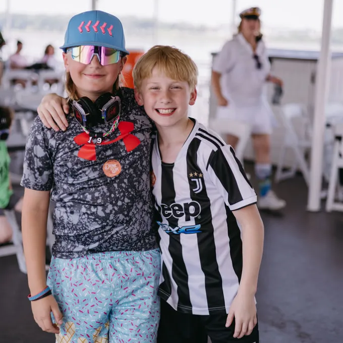 Silent Disco 2 Two kids smiling, one wearing sunglasses and a cap, the other in a striped sports jersey, indoors.