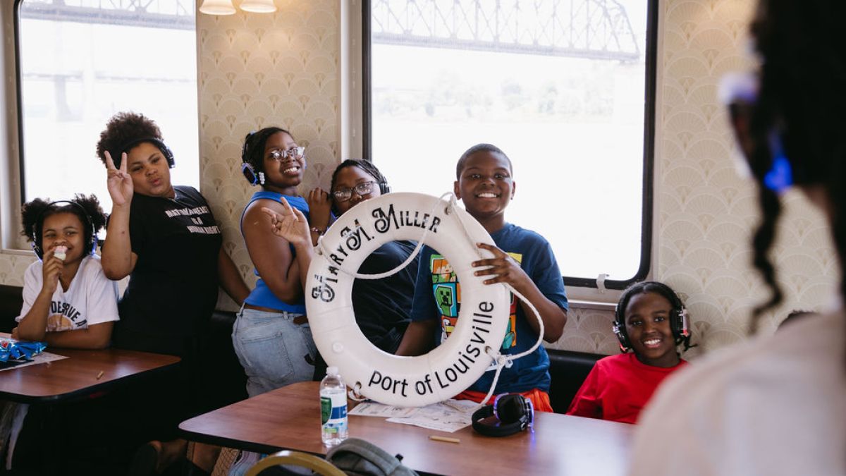 Group of kids on a boat posing with a life preserver labeled 'Port of Louisville.'