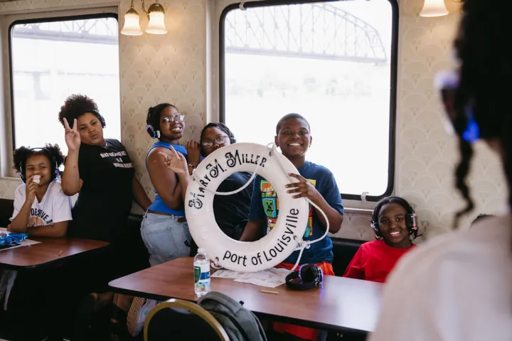 Group of kids on a boat posing with a life preserver labeled 'Port of Louisville.'