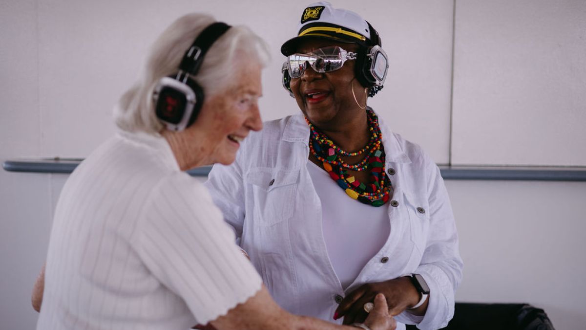 Two women wearing headphones, smiling and holding hands in a well-lit room.
