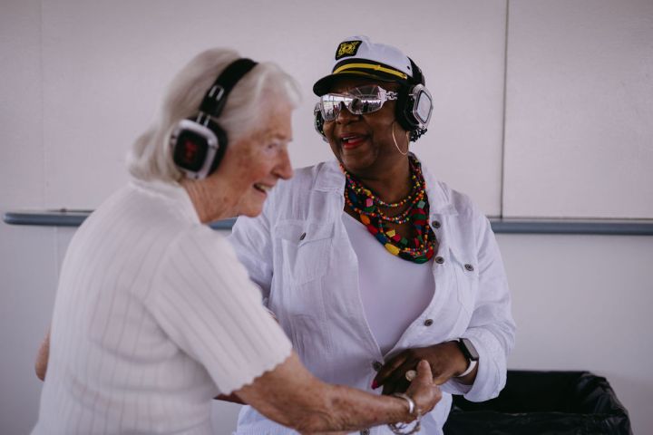 Two women wearing headphones, smiling and holding hands in a well-lit room.