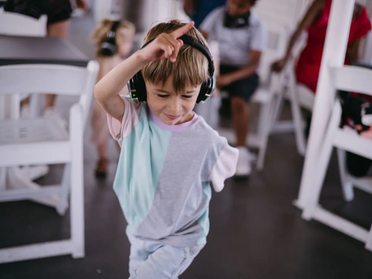 Young boy wearing headphones dances indoors, surrounded by chairs and people.