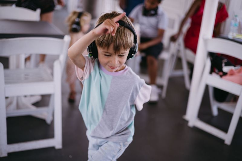 Young boy wearing headphones dances indoors, surrounded by chairs and people.