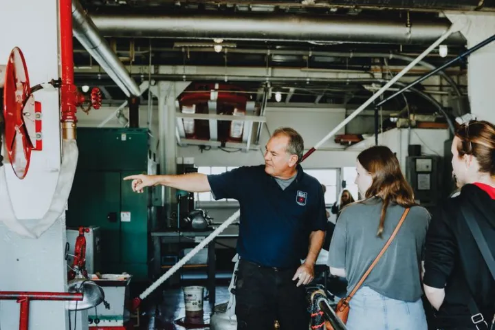 Man in dark blue shirt points at equipment while speaking to two people in an industrial setting.