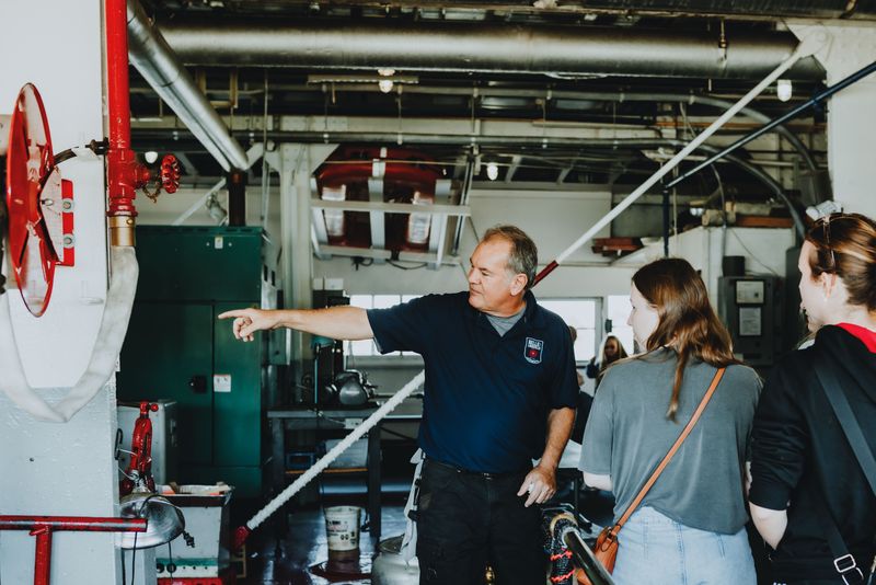 Man in dark blue shirt points at equipment while speaking to two people in an industrial setting.