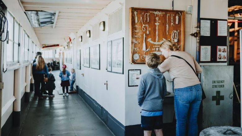 People in a corridor on a ship looking at a display of nautical knots on the wall.