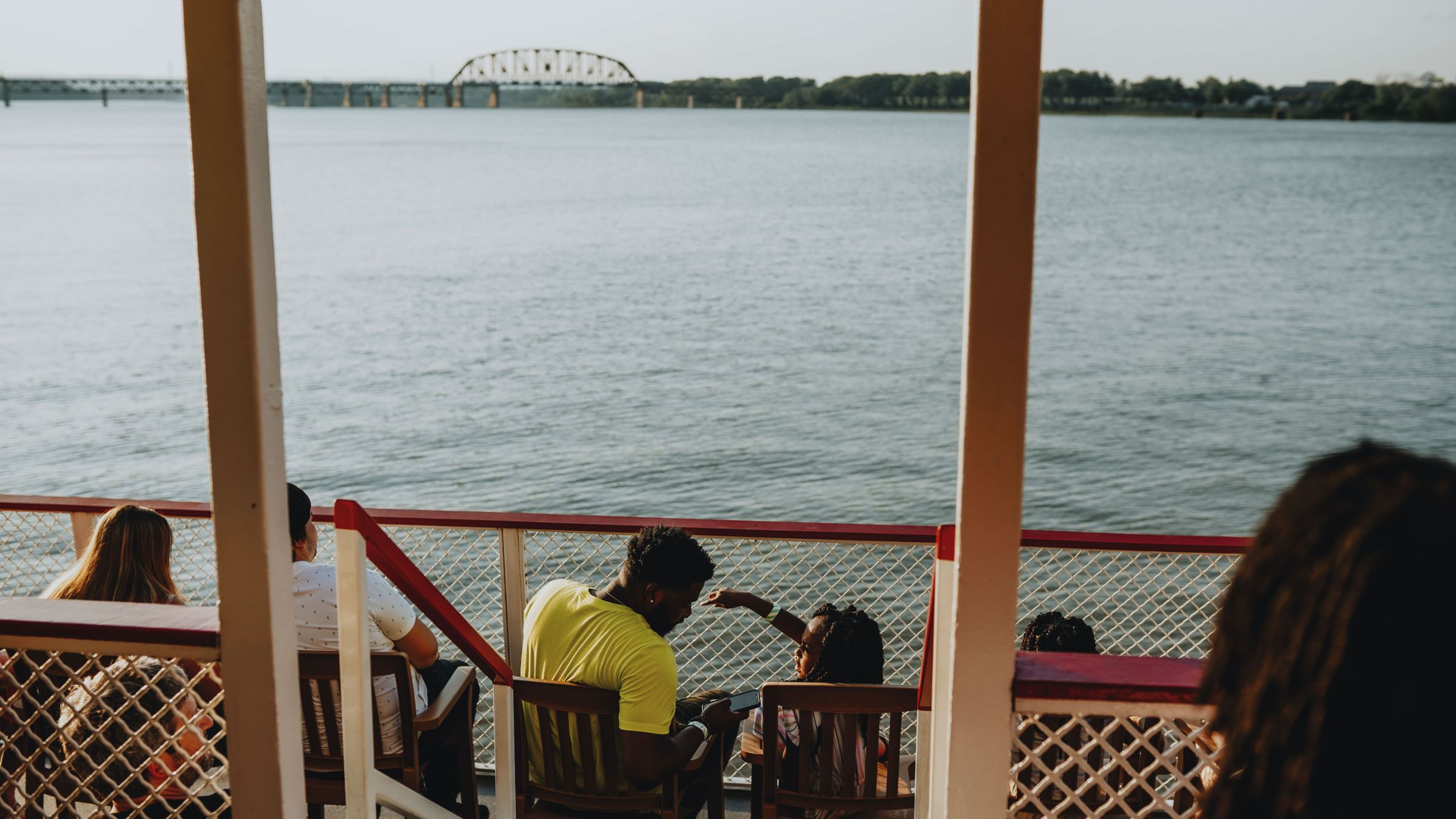 People sitting on a boat deck overlooking a river and distant bridge.