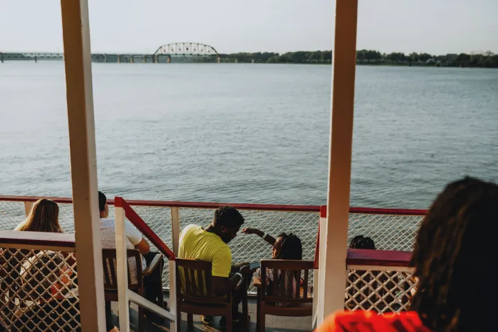 People sitting on a boat deck overlooking a river and distant bridge.