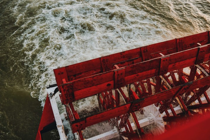 Red paddlewheel of a riverboat churning through water, viewed from above.