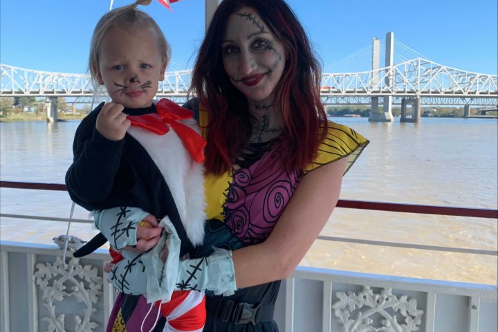 Two people in costume, one holding a child, with a bridge and river in the background on a sunny day.