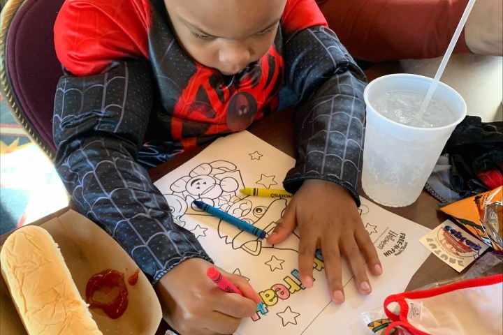 Child in costume coloring a picture with crayons at a table, next to a hot dog and drink.