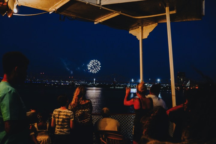 People on a boat watching fireworks over a river at night.