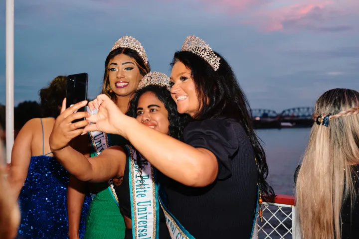 Three women wearing tiaras take a selfie on a boat at sunset.