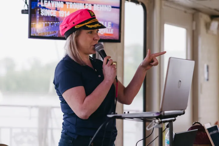 Woman with a pink captain's hat speaks into a microphone indoors with a laptop and audio mixer.