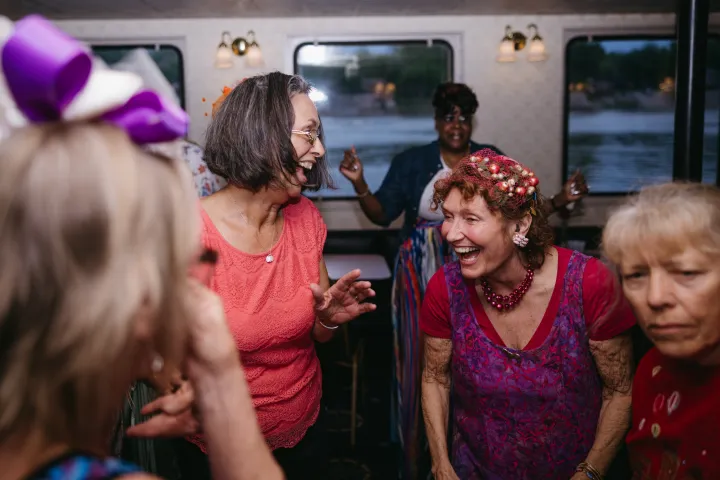Group of women laughing and talking on a boat with windows and lake view.