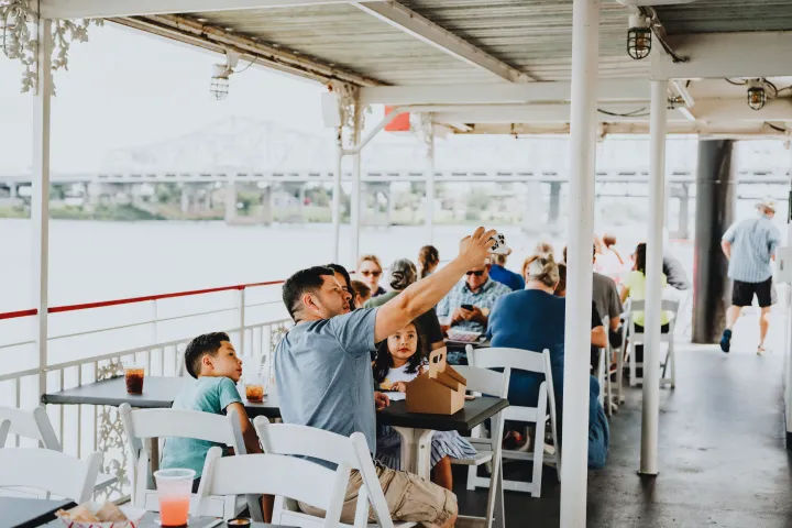 People sitting at tables on a riverboat, one taking a selfie, with a bridge in the background.