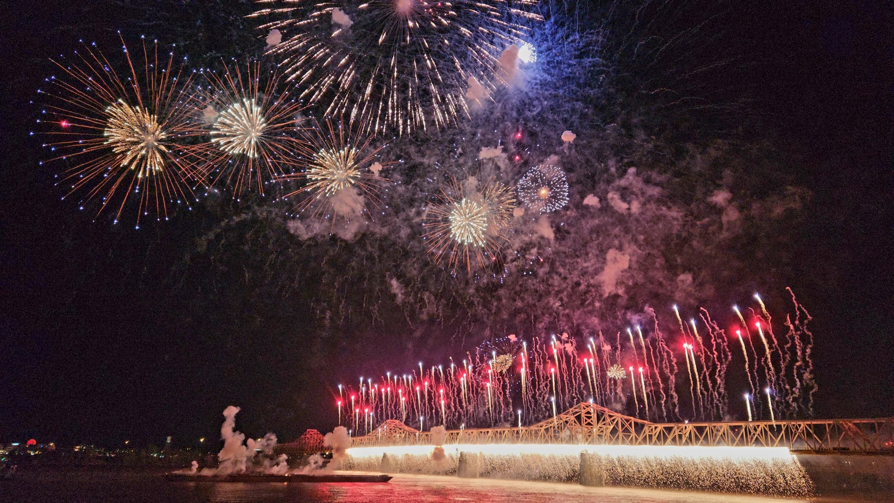 Colorful fireworks display over a bridge at night, with reflections on the water below.