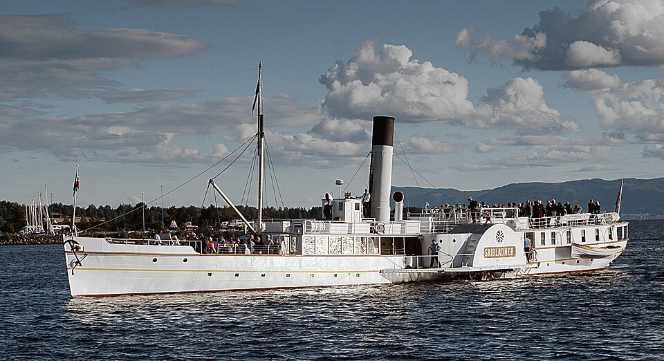 Historic paddle steamer with people on deck, sailing on a calm lake under a partly cloudy sky.
