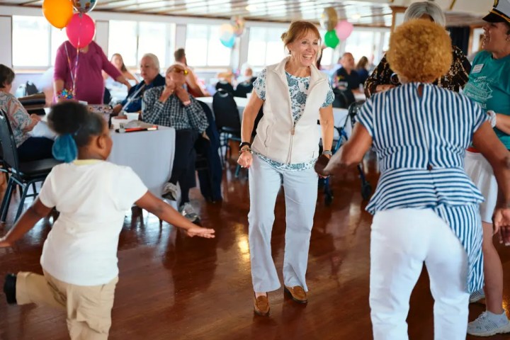 People of various ages dancing in a bright room with balloons and seated spectators.